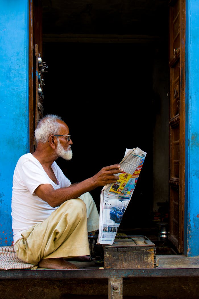 Senior man in white shirt reading a newspaper in a vibrant blue doorway in Bikaner, India.